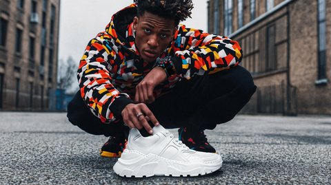 A young man crouches in an urban setting, wearing a bold, geometric-patterned hoodie with black pants. He holds a chunky all-white sneaker in focus, showcasing its sculpted sole and sleek design. His intense gaze and confident pose exude street-style energy, complemented by vibrant footwear details and accessories. The background features brick buildings and an industrial cityscape, adding depth and attitude to the shot.