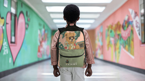 Young boy in pastel pink shirt walking down a colorful school hallway, wearing a ['LIT·TLE] HŪMN backpack with a stylish bear design. The vibrant murals and modern streetwear backpack create a trendy, youthful vibe.
