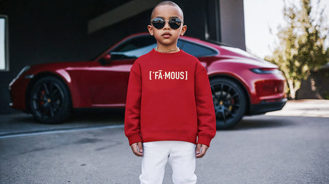 A stylish young boy wearing sunglasses, a red sweatshirt with the text "[FĀ·MOUS]" printed on the front, and white pants, standing confidently in front of a sleek red sports car parked in a modern outdoor setting