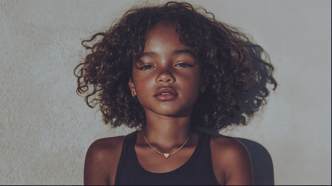 Young girl with curly hair wearing a delicate gold heart necklace, standing confidently against a textured wall, illuminated by soft natural light.