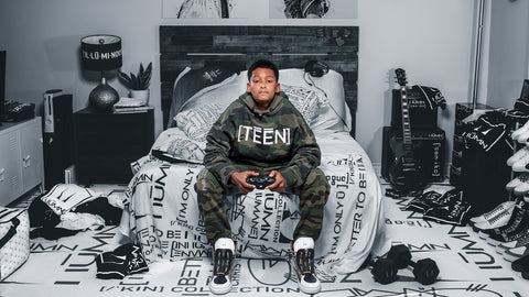 Teen boy in camo ['TEEN] hoodie sitting on a bed playing video games. The room is filled with modern streetwear, a guitar, and gaming gear. The bedding, rug, and decor display bold text and graphic designs, creating a trendy, urban, and laid-back atmosphere.