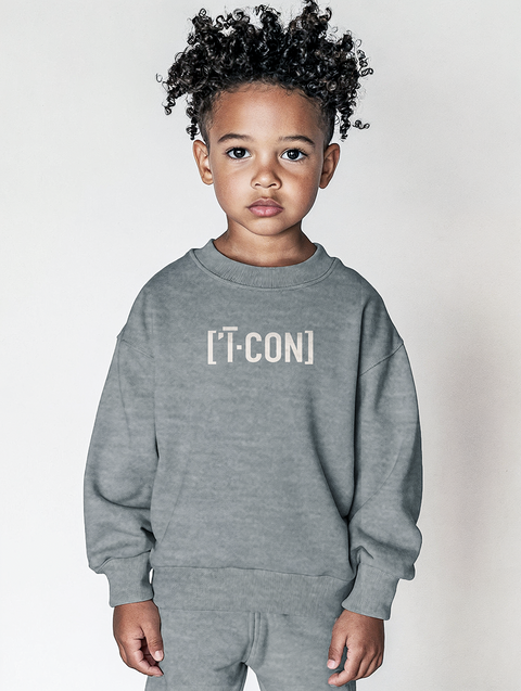 A young child wearing a soft grey sweatshirt and matching pants, with the text ICON printed on the chest in bold white. The minimalist outfit is cozy and modern, perfect for everyday wear.