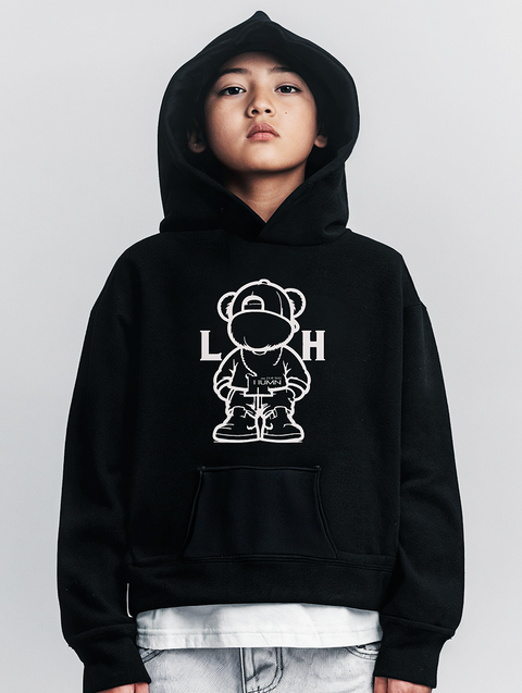 A child wearing a black hoodie featuring a graphic of a bear icon and the text LITTLE HUMN." The layered look with a white shirt peeking out gives a streetwear vibe that is bold and trendy.