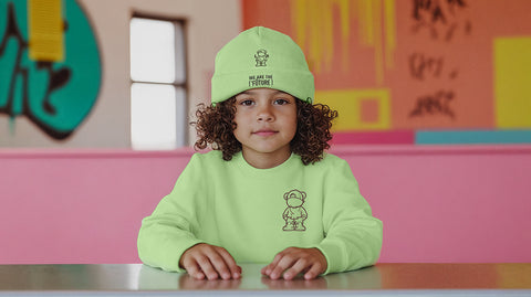 Young Latin child with curly hair sitting at a table wearing a neon green ['LIT·TLE] HŪMN sweatshirt and matching beanie with 'We Are The Future' text. The colorful background and stylish outfit create a trendy, modern look, embodying a youthful, confident vibe.