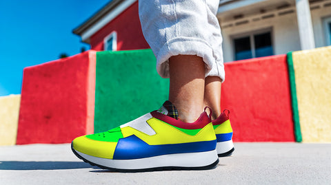 Close-up of a child wearing colorful ['LIT·TLE] HŪMN sneakers with bold yellow, blue, and green accents. The child's legs are shown against a vibrant outdoor backdrop featuring red, green, and yellow blocks, creating a playful and stylish look.