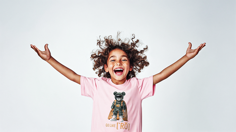 A joyful young child with curly hair wearing a light pink t-shirt featuring a graphic of a bear in a superhero outfit with the text GO LIKE RO. The child has arms raised in excitement, capturing the playful and empowering vibe of the design. The clean white background emphasizes the vibrant colors and energy of the t-shirt.
