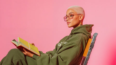 Stylish person with short blonde hair, glasses, and star tattoo, sitting on a chair wearing an olive green ['LIT·TLE] HŪMN hoodie while reading a book. The pink background and trendy outfit create a modern, confident look with an intellectual vibe.