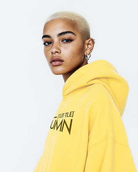 A confident young woman with a buzz cut wearing a bright yellow hoodie featuring the LITTLE HUMN logo in bold print. The design adds a pop of color and a fun, stylish edge to casual wear.