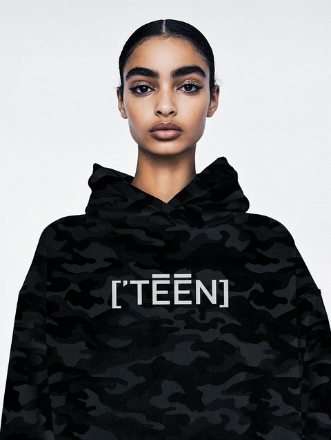 A confident young woman wearing a black camouflage hoodie with the text TEEN printed in bold white letters across the chest. The hoodie combines streetwear style with minimalistic design, perfect for a bold and modern look.