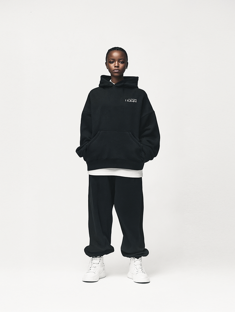 A young woman wearing a black oversized hoodie with the LITTLE HUMN logo on the chest, paired with black sweatpants and white high-top sneakers. The minimalistic, monochrome outfit exudes a relaxed, streetwear-inspired aesthetic, perfect for casual and comfortable wear.